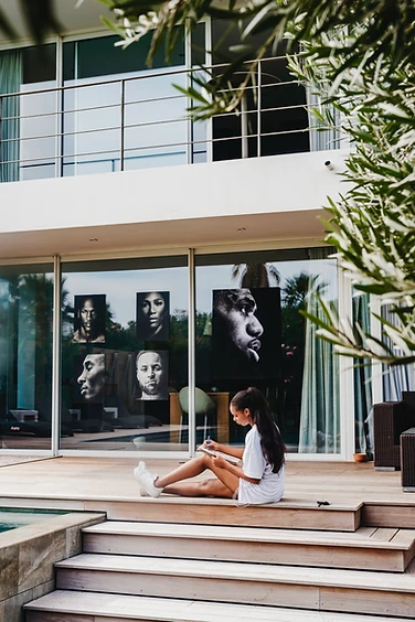Anaïa sketching on a deck, portraits of athletes visible through the glass doors