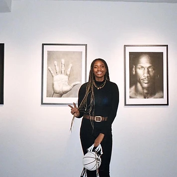 Anaïa in a gallery between charcoal portraits, holding a basketball handbag