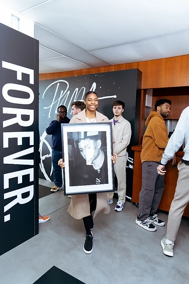 Anaïa holding a framed portrait, standing beside a FOREVER. panel