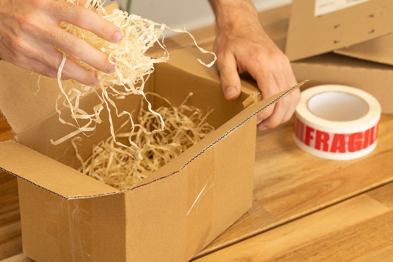 A person filling a cardboard parcel box with wood wool cushioning beside a roll of fragile tape.