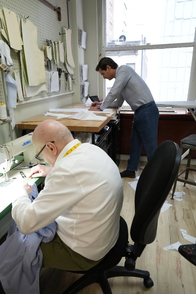 A tailor at the sewing machine, with a second tailor marking a pattern at the workbench