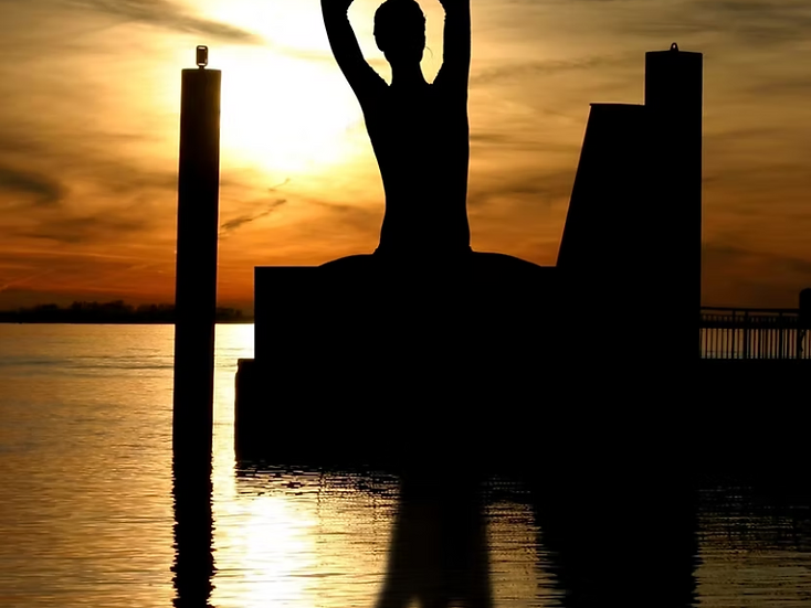 Silhouette practicing yoga beside the water at sunset