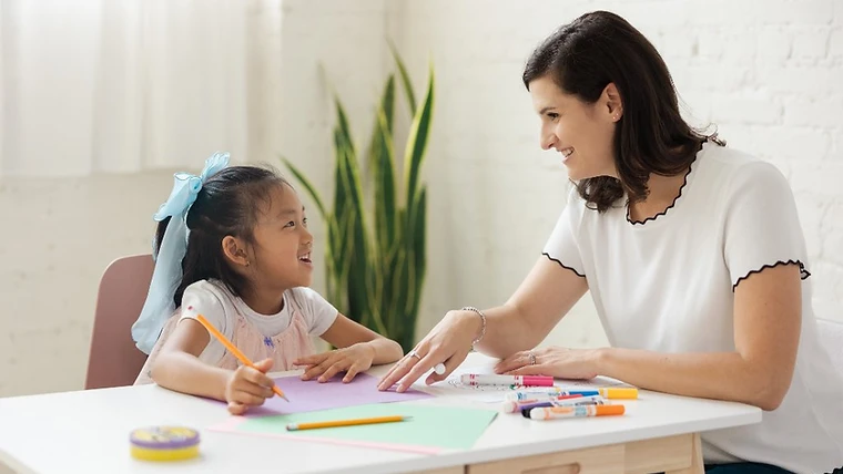 A woman smiling with a young girl during an art activity at a white table