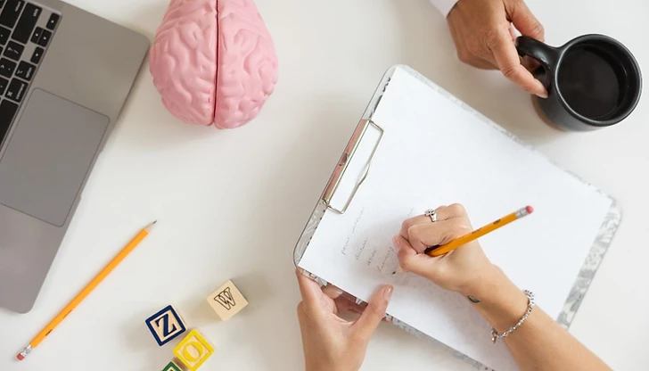 Hands writing on a clipboard with a brain model, alphabet blocks, and a laptop on a white surface