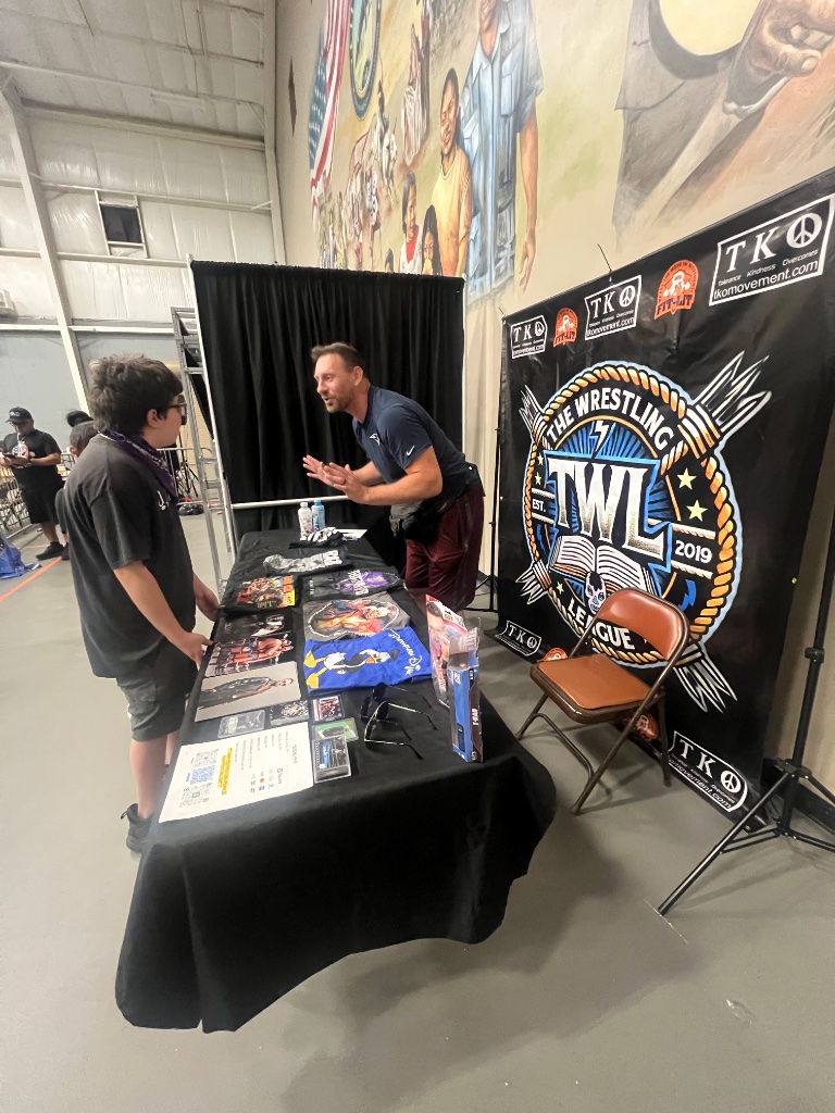 An attendee chats with a wrestler at The Wrestling League booth, surrounded by merch and the Pharr Pride mural.