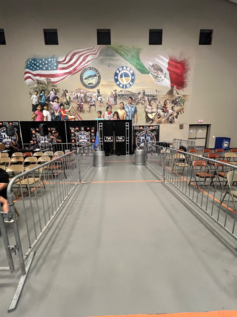 The entrance aisle lined with barricades and folding chairs, set against the Pharr Pride mural before the action begins.