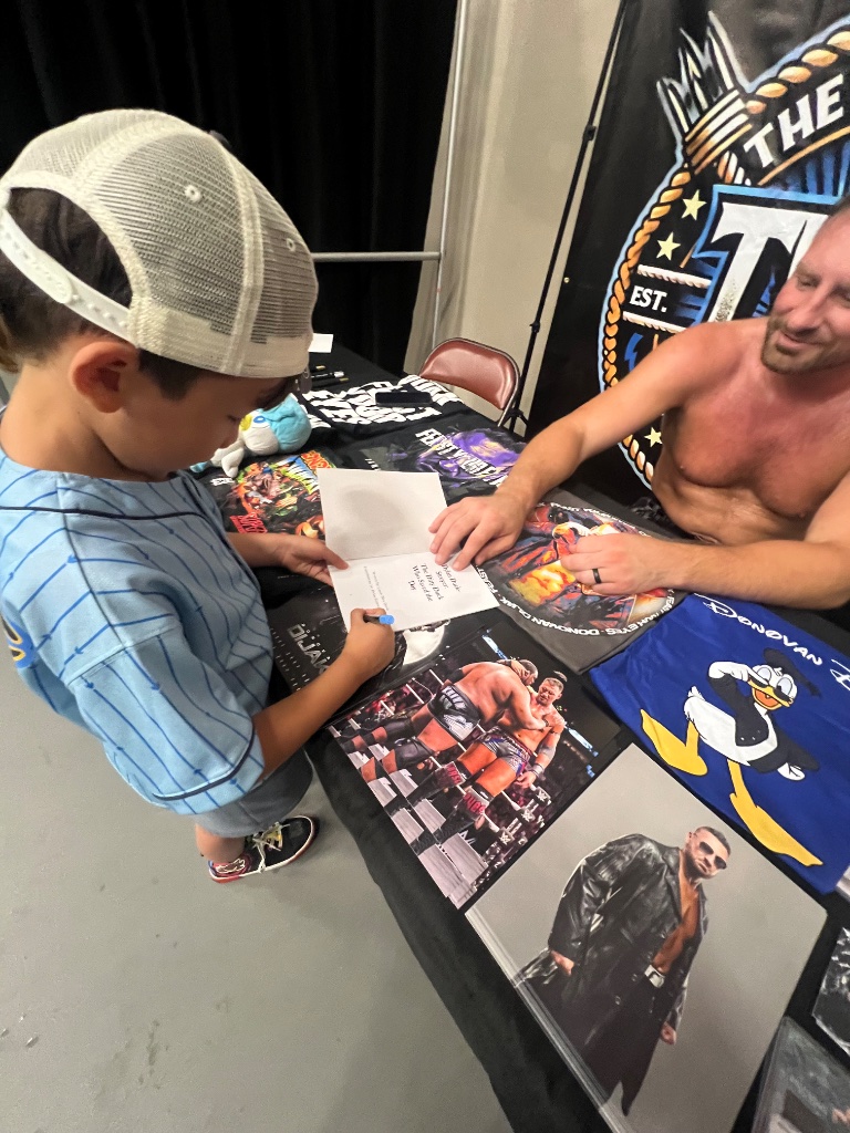 A young fan signs an autograph card alongside wrestler Donovan Dijak at the Lucha Libros 2025 meet-and-greet.