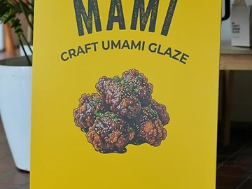 Yellow MAMI launch signage reading "MAMI Craft Umami Glaze" with product artwork, displayed beside stacked MAMI boxes credited to Sauce Labs, Singapore