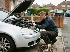 Auto electrician testing wiring in a car's engine bay on a residential driveway