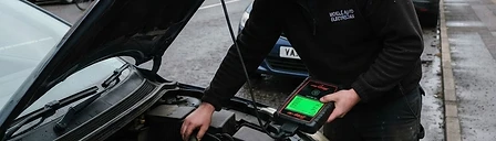 Auto electrician diagnosing a car's electrical system at the roadside with the bonnet open