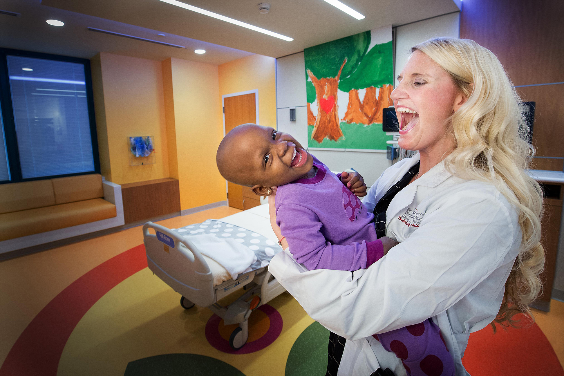 A smiling St. Jude patient being held by a St. Jude medical professional in a patient room