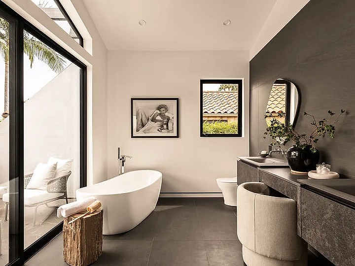 A primary bath with a white freestanding tub beside a glass wall to a private terrace, dark stone walls and a floating stone vanity, at La Jolla North in West Hollywood, by Apel Design