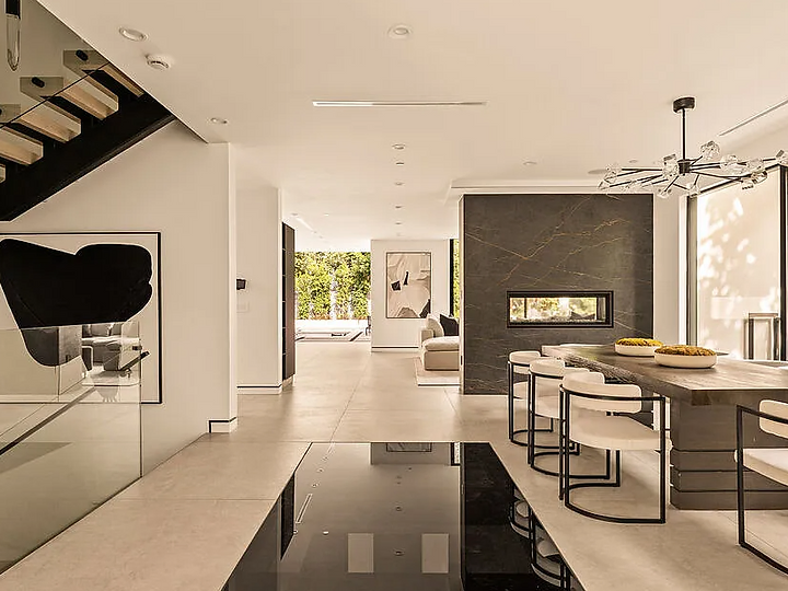 A dining room under a branching glass chandelier, divided from the living room by a double-sided stone fireplace, at La Jolla North in West Hollywood, by Apel Design