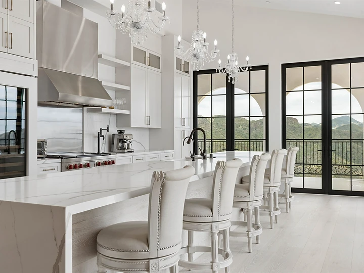 White kitchen at Oya with a marble island, upholstered stools and steel-framed doors to the terrace