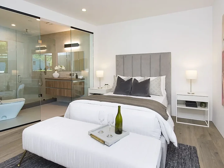 A tall upholstered headboard and a bench at the bed's foot, beside a glass-walled bath, an interior by Apel Design