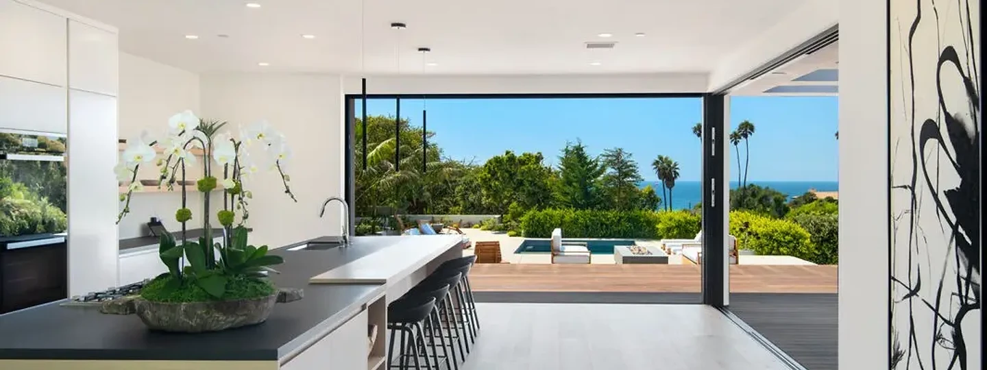 Kitchen island with a dark counter leading to an open glass wall, a pool and an ocean view, an interior by Apel Design