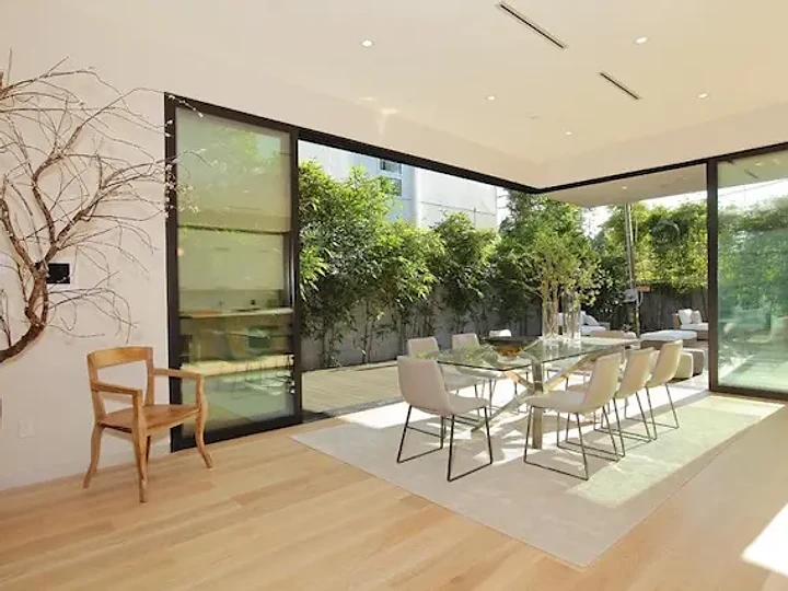 A dining table beside black-framed glass doors slid open to a green garden, an interior by Apel Design