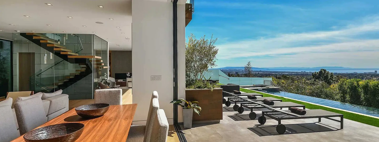 A timber dining table beside a wide opening onto a pale paved terrace and view, an interior by Apel Design