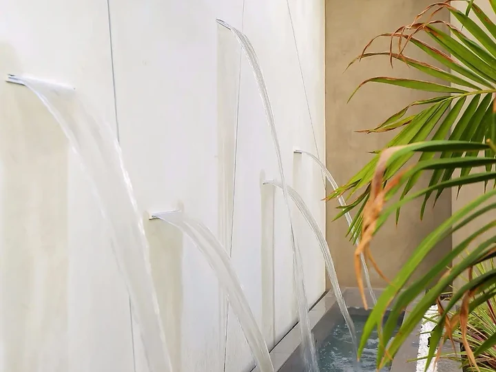Streams of water falling from a smooth white wall into a narrow pool beside palms, an interior by Apel Design