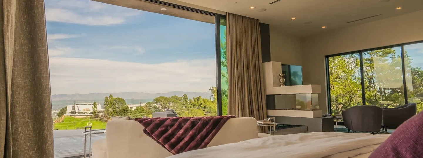 A primary suite with full-height glass and tall drapery framing hills and sky, an interior by Apel Design