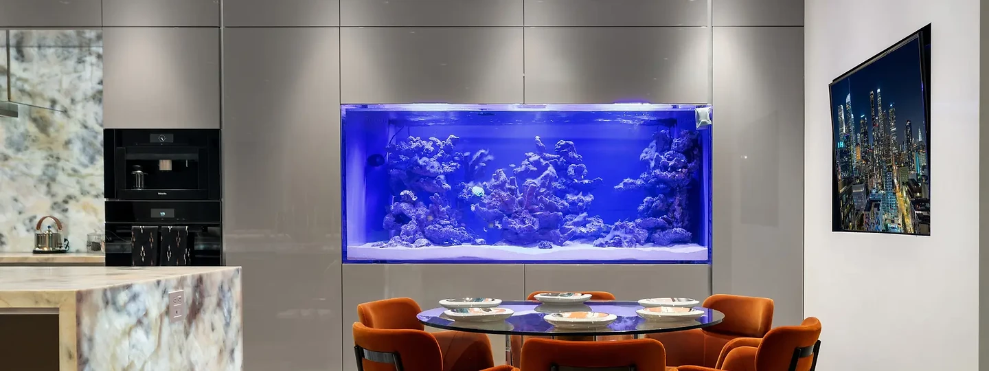 Orange dining chairs beside a waterfall stone island and glossy gray cabinetry framing an aquarium, an interior by Apel Design