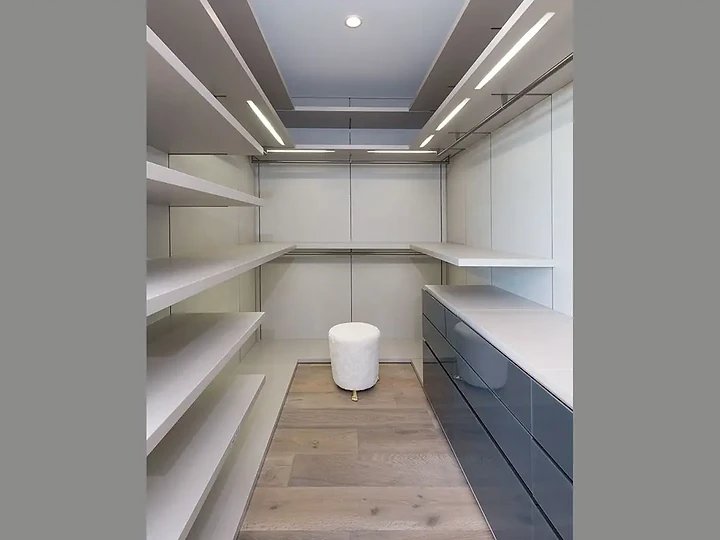 A walk-in closet of open white shelving under linear light slots in a stepped ceiling, an interior by Apel Design