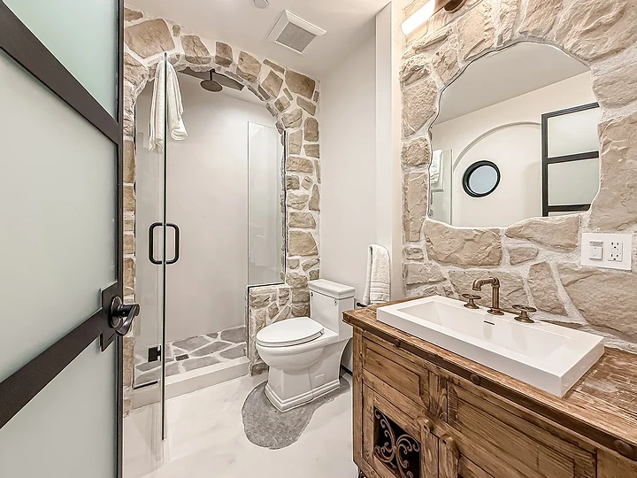 Guest bath with a stone-framed mirror, a carved wood vanity and a glass shower, at Mintcello in Malibu, by Apel Design