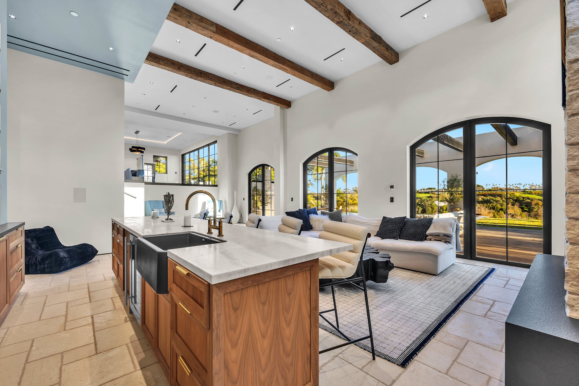 The kitchen island and the great room beyond, under timber beams, Merritt Estate in Malibu by Apel Design