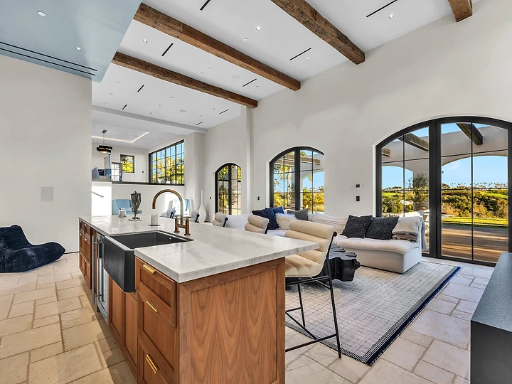 The kitchen island opens to the great room under timber beams, at Merritt Estate in Malibu, by Apel Design