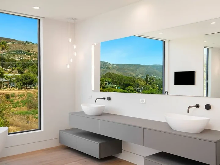 Floating grey vanity with two vessel basins beside a tall window onto green hills, an interior by Apel Design