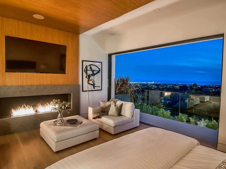 From the bed, a linear fireplace and a glass wall opening to evening lights, an interior by Apel Design
