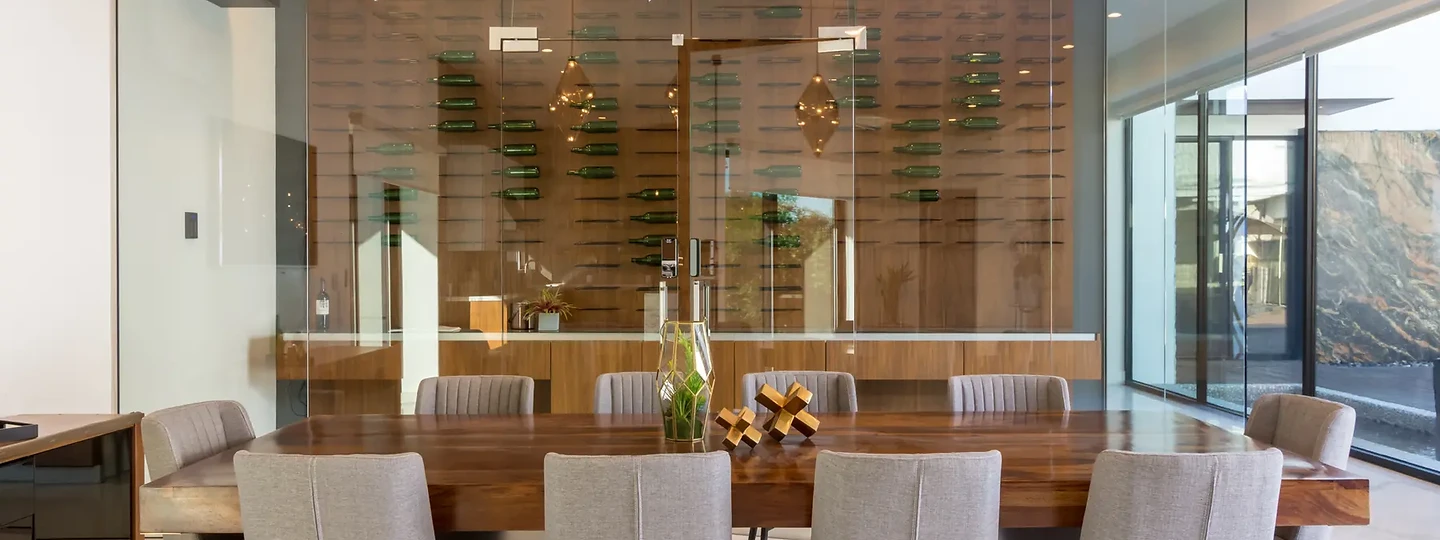 Three faceted pendants over a long timber table before a glass-walled wine room, an interior by Apel Design