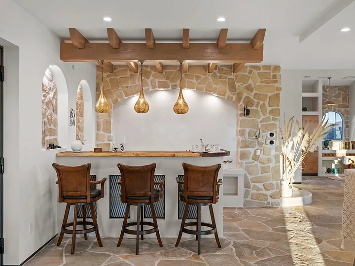 Stone bar with three stools under a timber trellis and pendant lights, at Mintcello in Malibu, by Apel Design