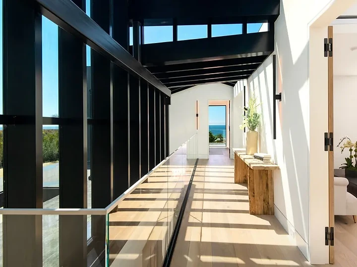 A long hall of black-framed glass under dark beams, ending at an ocean view, an interior by Apel Design