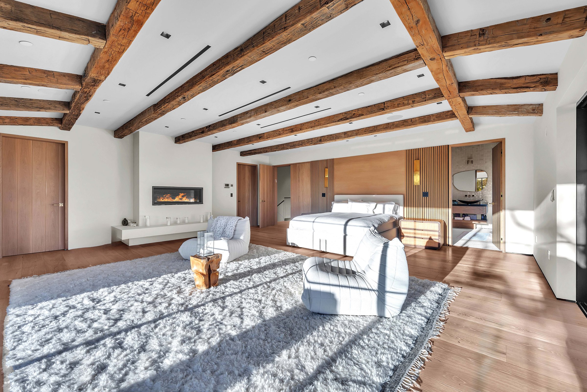The primary suite, with a linear fireplace under timber beams, Merritt Estate in Malibu by Apel Design