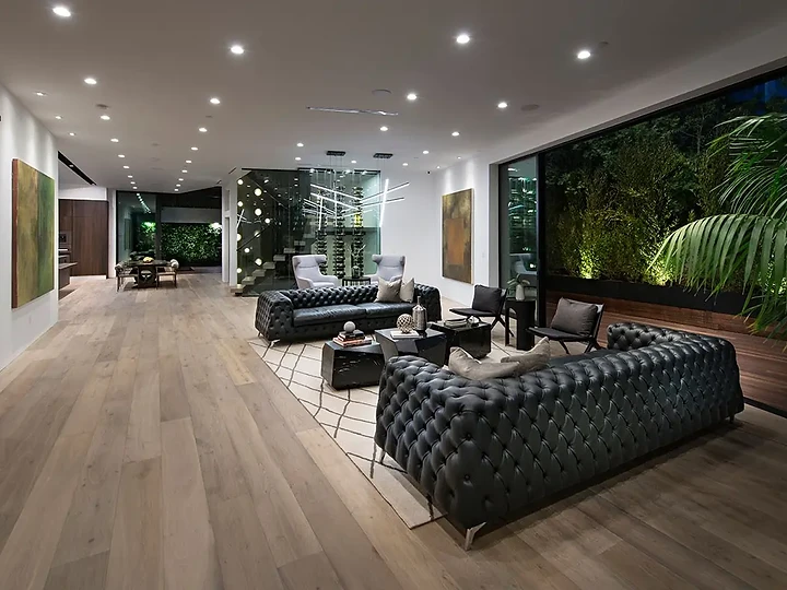 Black tufted sofas under recessed lights beside a glass wall onto a lit garden, an interior by Apel Design