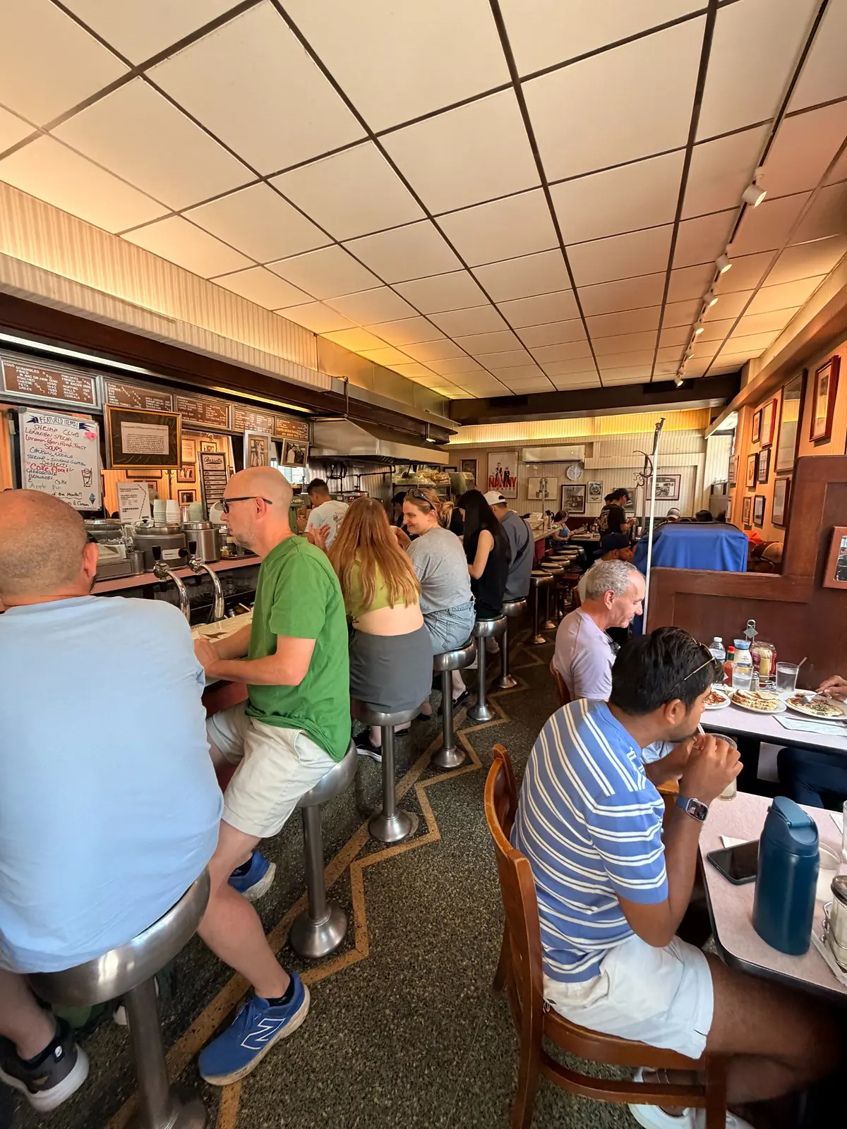 Wide interior view of Lexington Candy Shop, with customers seated on metal counter stools and at closely arranged tables.