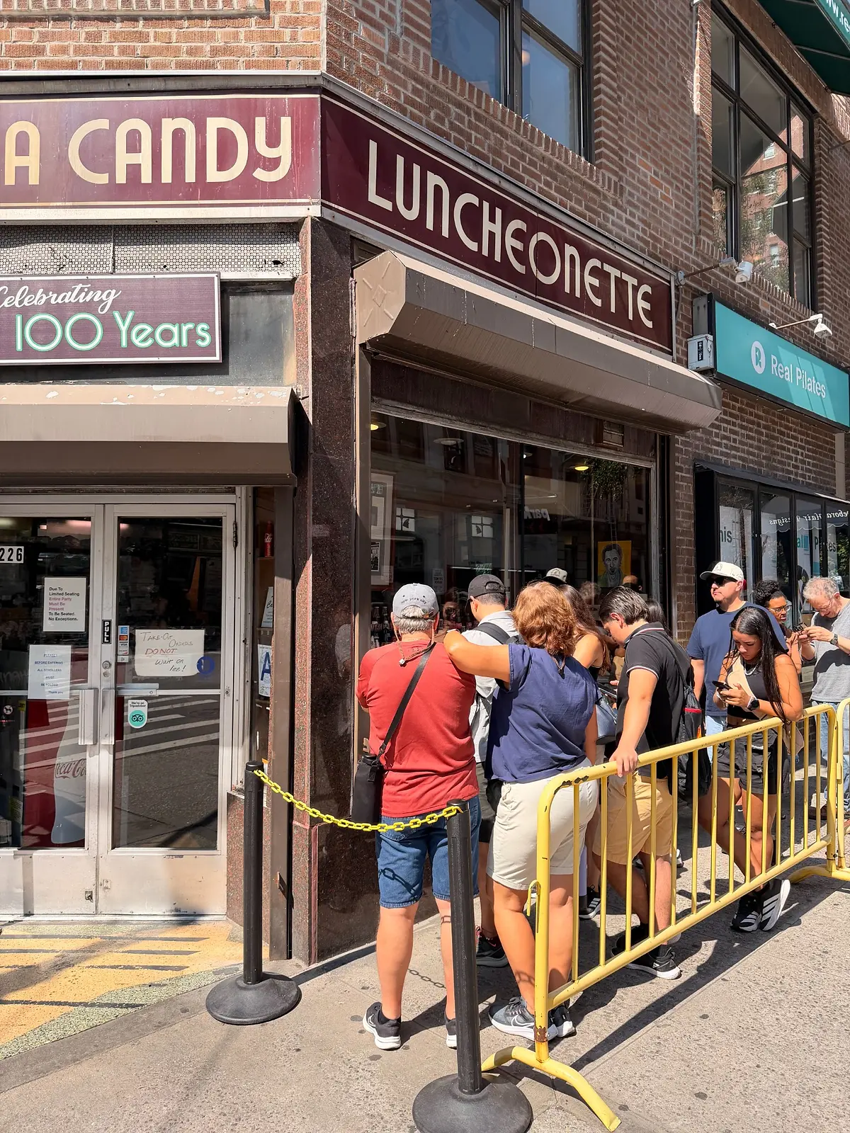 Customers waiting behind a yellow barrier outside the luncheonette entrance beneath the vintage LUNCHEONETTE sign.