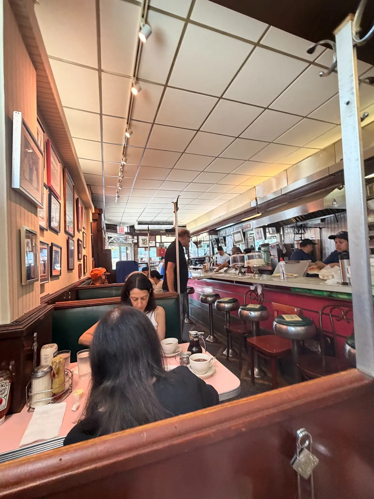 View through Lexington Candy Shop’s narrow dining room, showing green booths, pink tables, counter stools, and the open kitchen.