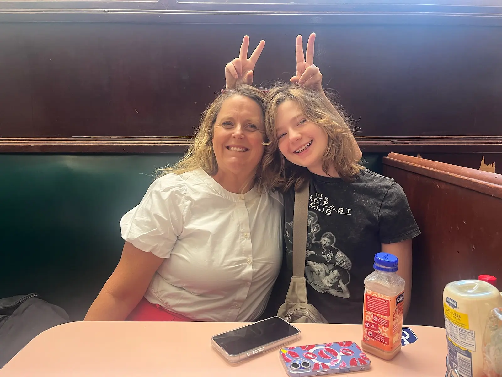 Chrissy and Olivia smiling together in a green booth at Lexington Candy Shop while hands behind them make bunny ears over their heads.