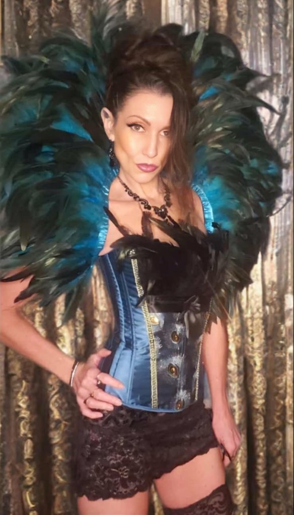 Empress Steele in a blue satin corset and feather collar against a gold curtain