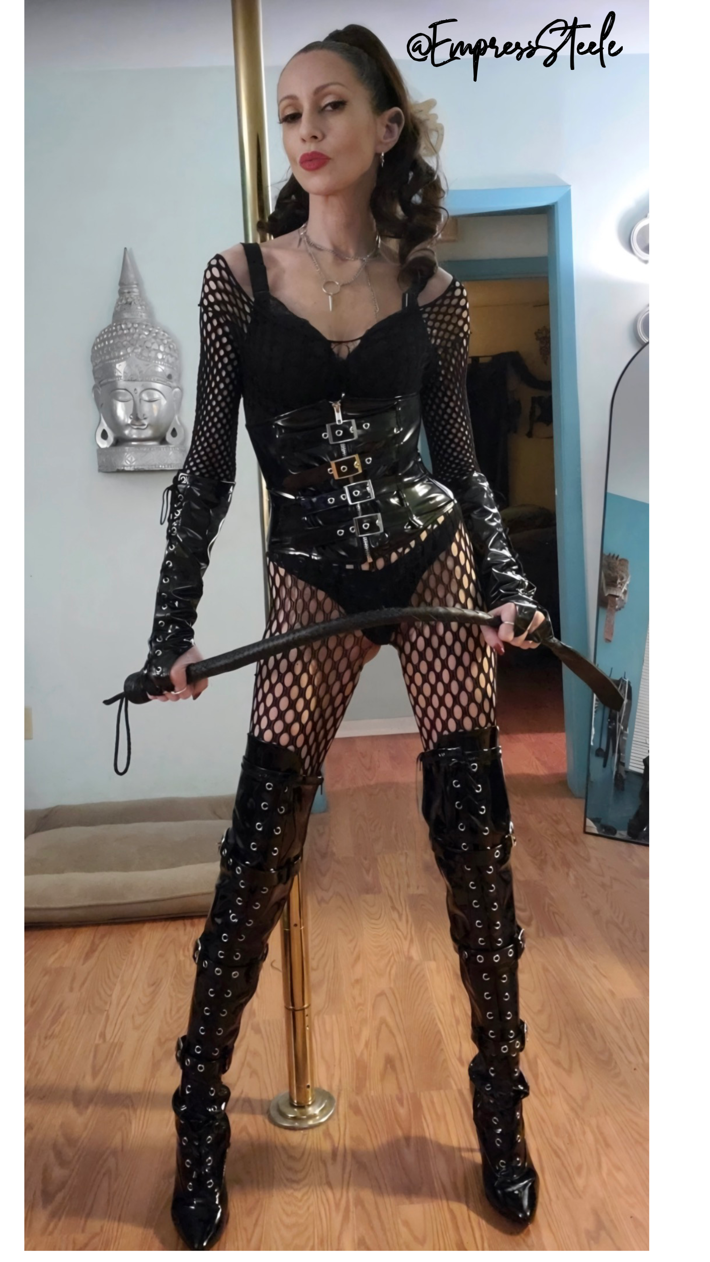 Empress Steele in a black patent corset, fishnet top, and thigh-high buckle boots holding a whip