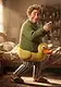 A friendly man with curly brown hair wearing a green sweater and a yellow duck floatie around his waist, standing in a cozy bedroom while holding a colorful sock.