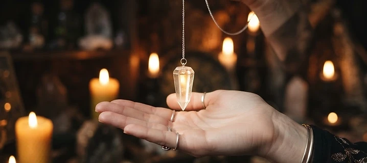 A crystal pendulum on a silver chain held still above an open hand in dim candlelight