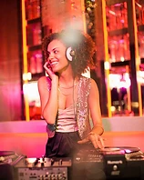 Female DJ in a sequined vest smiling behind the decks under magenta and amber light