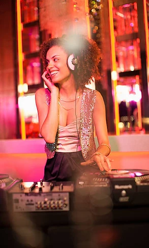 Female DJ in a sequined vest smiling behind Numark decks under magenta and amber club light