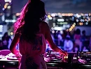 Female DJ seen from behind, mixing on a rooftop terrace bathed in magenta light