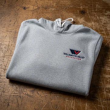 Grey hoodie with Willamette Valley Excavating embroidered logo on rustic wood surface