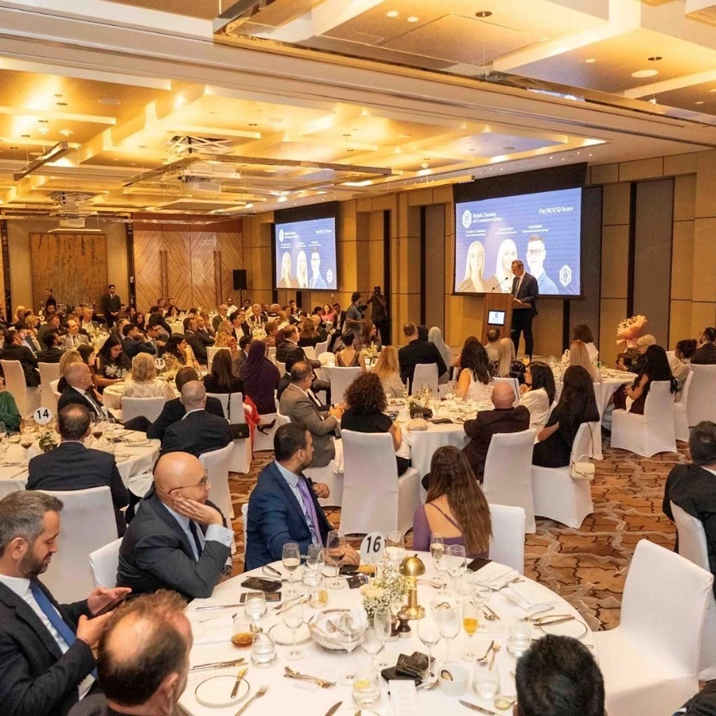 Guests seated at round tables in a chandelier-lit ballroom during the British Chamber of Commerce Qatar Celebration Evening at Park Hyatt Doha, with a speaker at a podium and British Chamber of Commerce Qatar branding on large screens behind the stage.