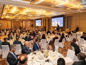 Guests seated at round tables in a chandelier-lit ballroom during the British Chamber of Commerce Qatar Celebration Evening at Park Hyatt Doha, with a speaker at a podium and British Chamber of Commerce Qatar branding on large screens behind the stage.